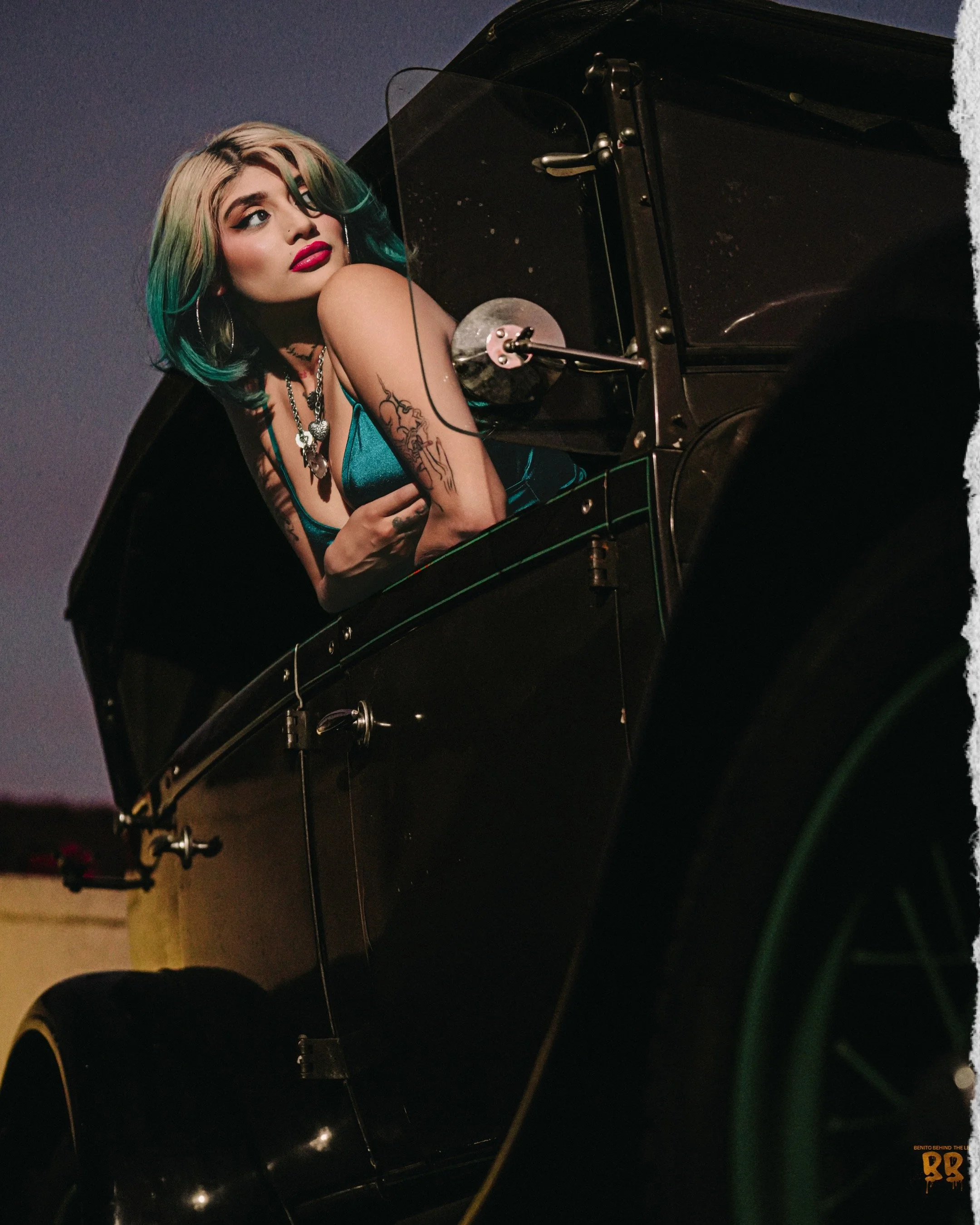 A woman with multi-colored hair (blonde with green tips), bold makeup, and tattoos on her arm, sitting at a black grand piano, looking to the side, wearing jewelry, in a dimly lit setting.