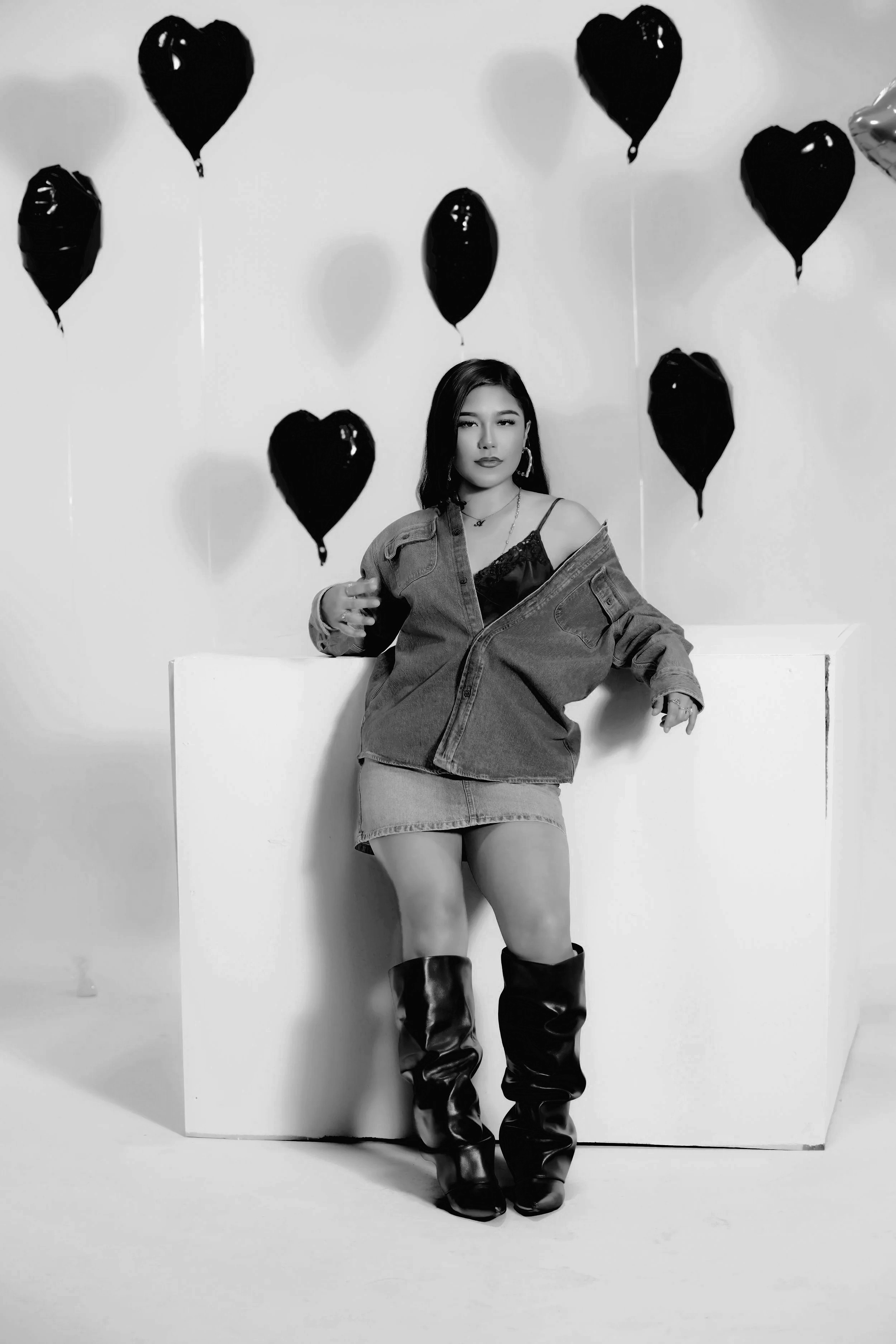 Black and white photo of a woman posing in front of a white background with black heart-shaped balloons. She is wearing a denim jacket, a lace top, a short skirt, and knee-high boots, with jewelry accessories.