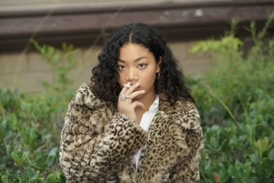 A woman with curly black hair wearing a leopard print coat, sitting outdoors with green foliage and wooden stairs in the background.Photographer, for hire, Oceanside, Carlsbad, San Diego, Orange County, Fashion, Portraits, Moments, Lifestyle, Events,