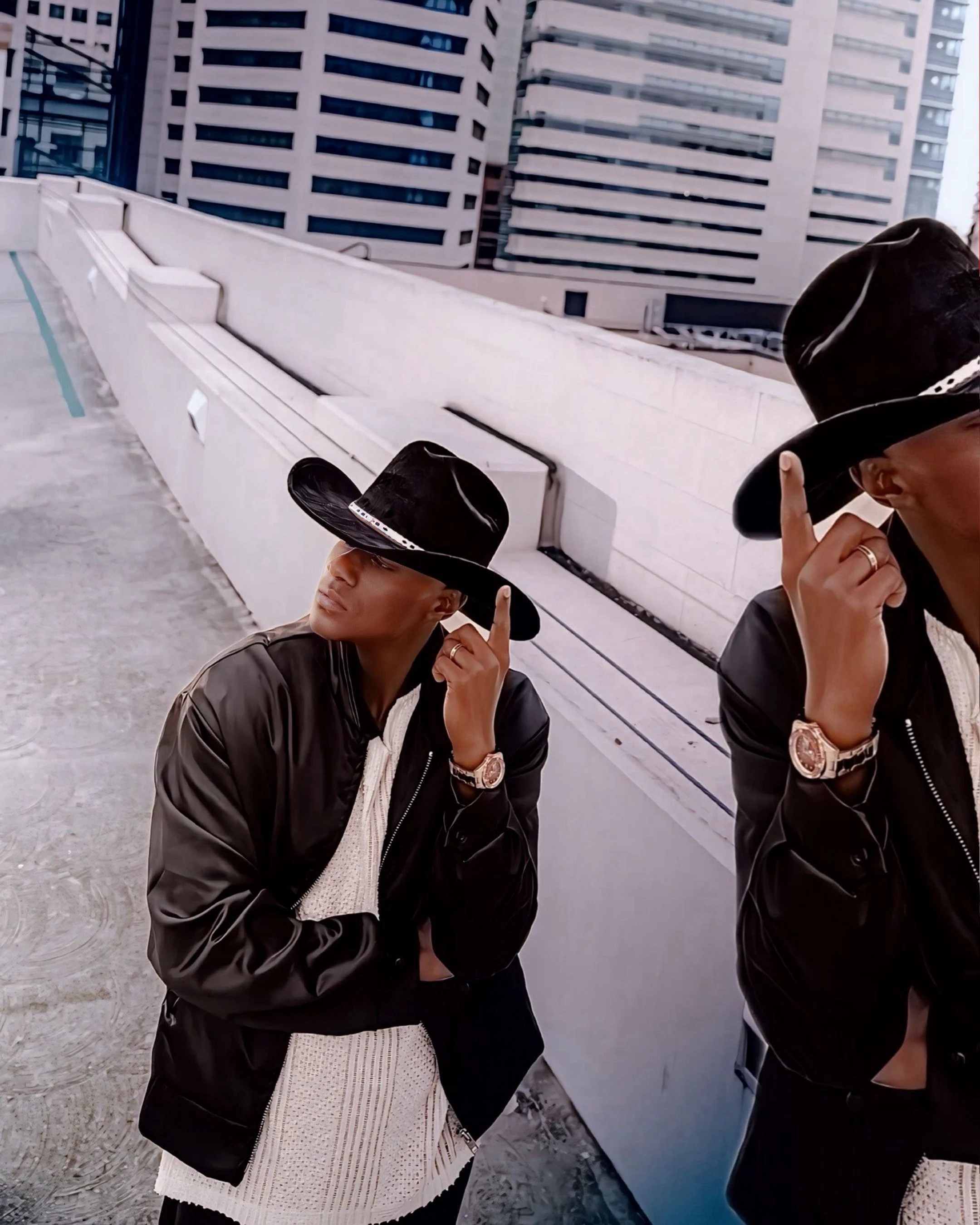 A woman is on a rooftop in an urban area, wearing a black wide-brimmed hat and a black leather jacket. The photo is taken from a low angle, capturing her face partially while she is adjusting her hat with her right hand. Behind her, there are tall modern buildings with numerous windows. The sky is overcast.