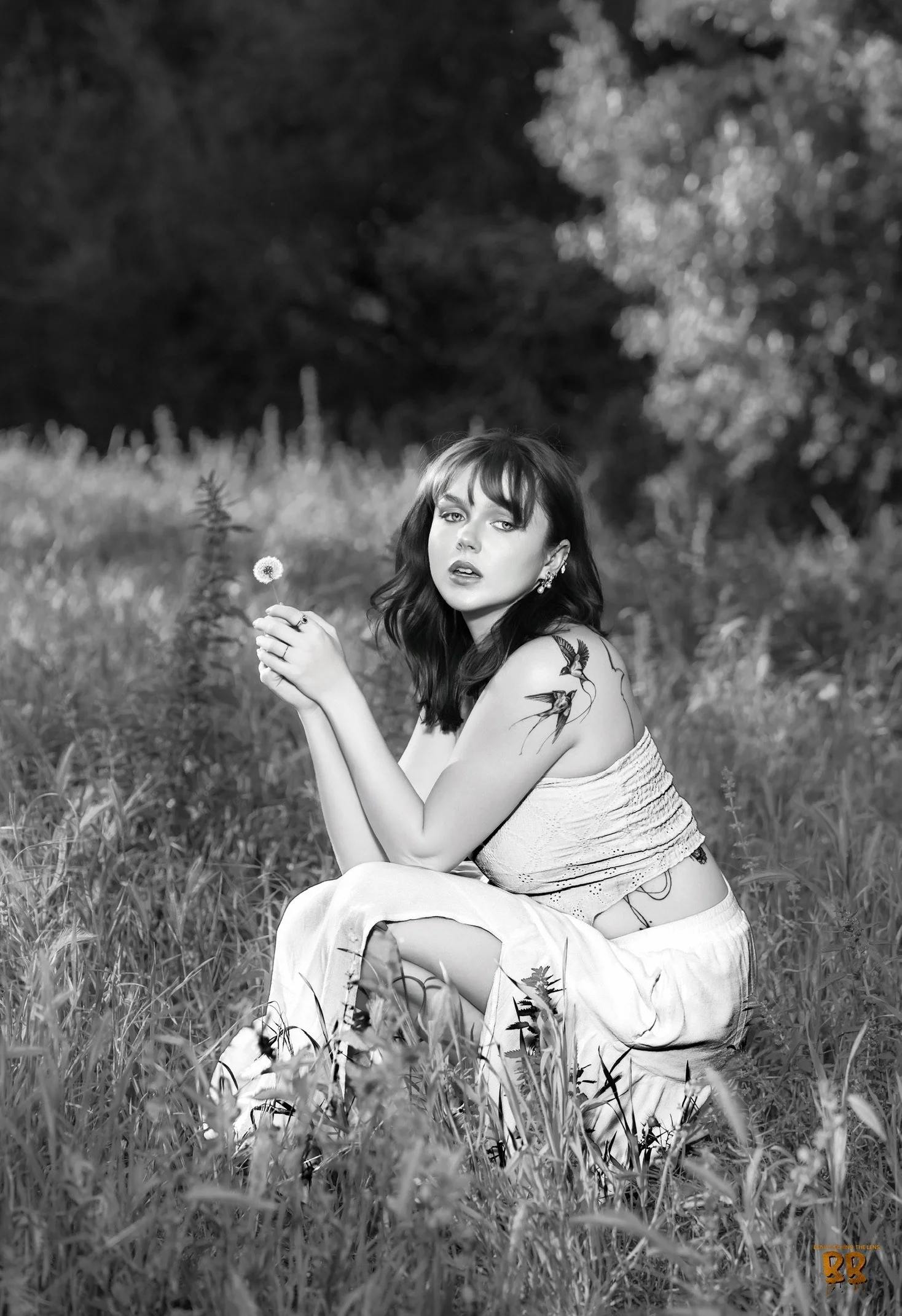 A woman with dark hair and tattoos on her arm sitting in a grassy field, holding a dandelion, with trees in the background, in black and white.