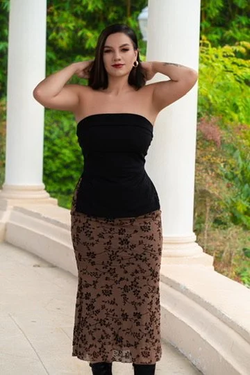 A woman standing outdoors on a concrete pathway, surrounded by greenery and white columns, wearing a strapless black top and a long patterned skirt.Photographer, for hire, Oceanside, Carlsbad, San Diego, Orange County, Fashion, Portraits, Moments, Li