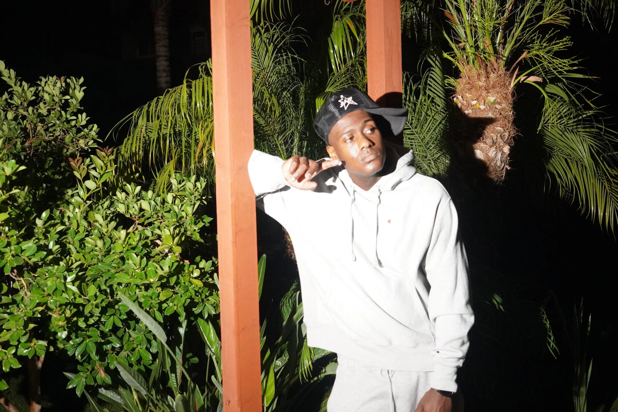 Young man wearing a black cap with a star and bubble logo, a white hoodie, and light-colored pants standing outdoors at night surrounded by lush green plants and palm trees.