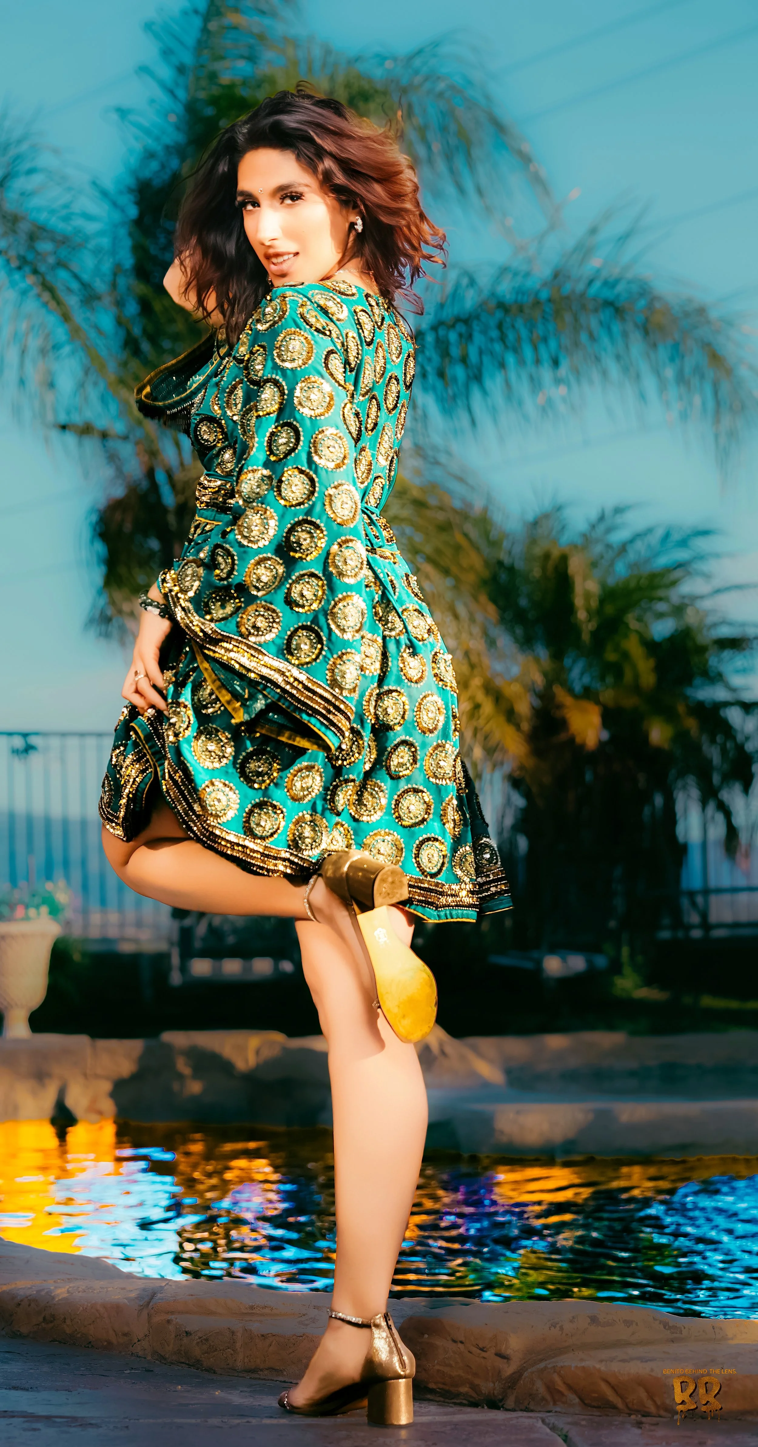 Woman in a colorful, patterned dress standing near a pool with palm trees in the background during sunset.Oceanside engagement photographer, beach photoshoot Oceanside, couples portraits SoCal, lifestyle photographer.high-end event media, cinematic.