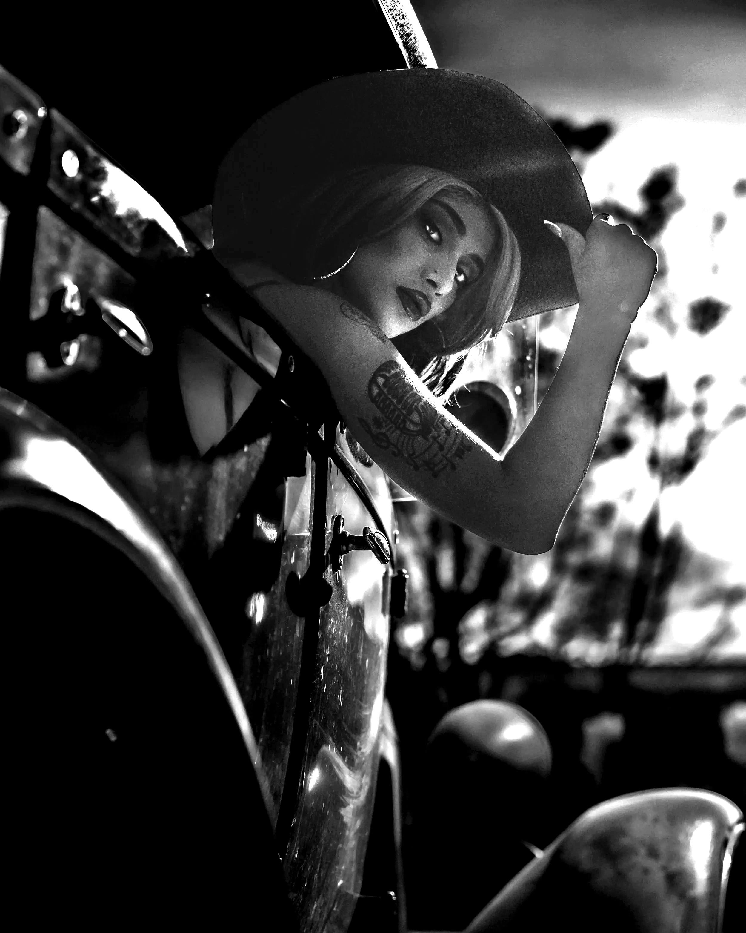Black and white photo of a woman wearing a wide-brimmed hat, lying on a motorcycle with her arm stretched above her head, looking at the camera. Modeling, Portraits, Images, Photography, Creative, Vision.
