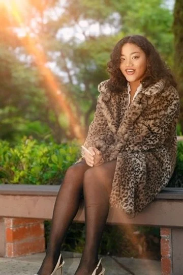 Young woman sitting on a park bench wearing a leopard print coat and black tights, with trees and sunlight in the background.Photographer, for hire, Oceanside, Carlsbad, San Diego, Orange County, Fashion, Portraits, Moments, Lifestyle, Events, Brandi