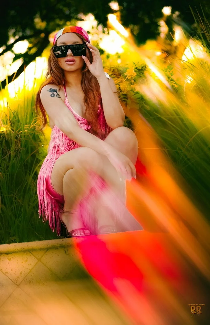 A young woman in a pink dress and sunglasses sits in a grassy outdoor setting during sunset, with warm sunlight filtering through trees.