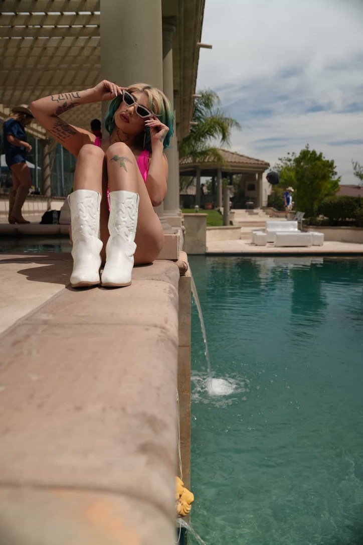 A woman with blonde hair and tattoos sitting on the edge of a swimming pool, wearing white cowboy boots, pink swimsuit, and sunglasses, with a pool and palm trees in the background.