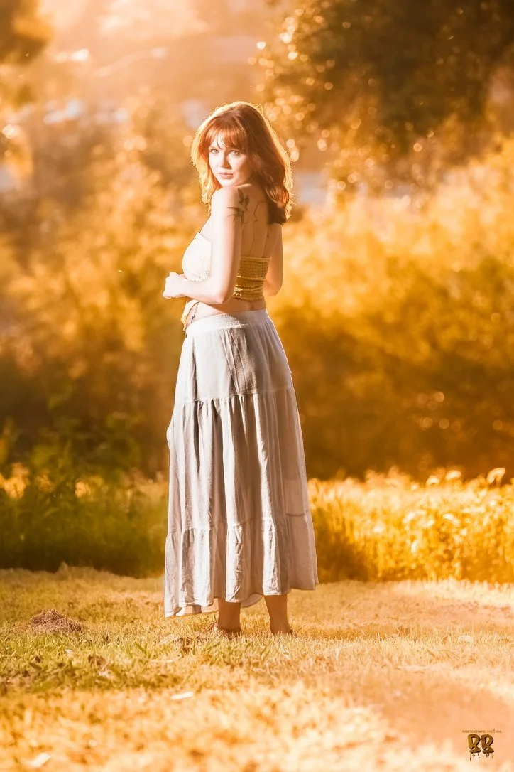 A woman with red hair standing outdoors during sunset, wearing a white crop top and a long, flowing skirt, with trees in the background.