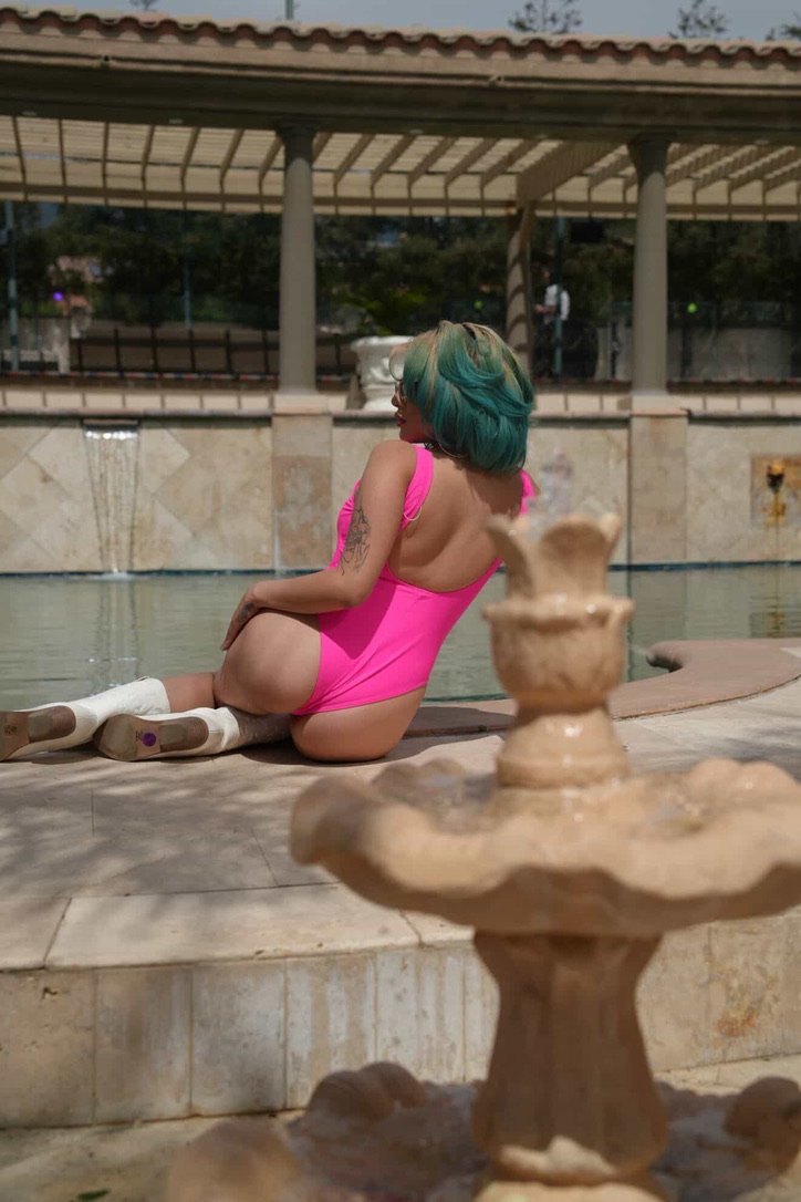 A woman with teal hair sits by a fountain near a pool, wearing a bright pink swimsuit, with one leg outstretched and the other bent. She is looking to the side, showing her tattoos on her arm. The background features a pergola and pool area.