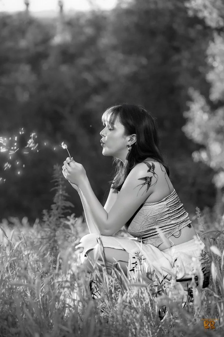 A woman with dark hair, sitting with legs crossed in a field, holding a sparkler, with a tattoo on her right arm, wearing a strapless top and loose pants, in a black and white photo
