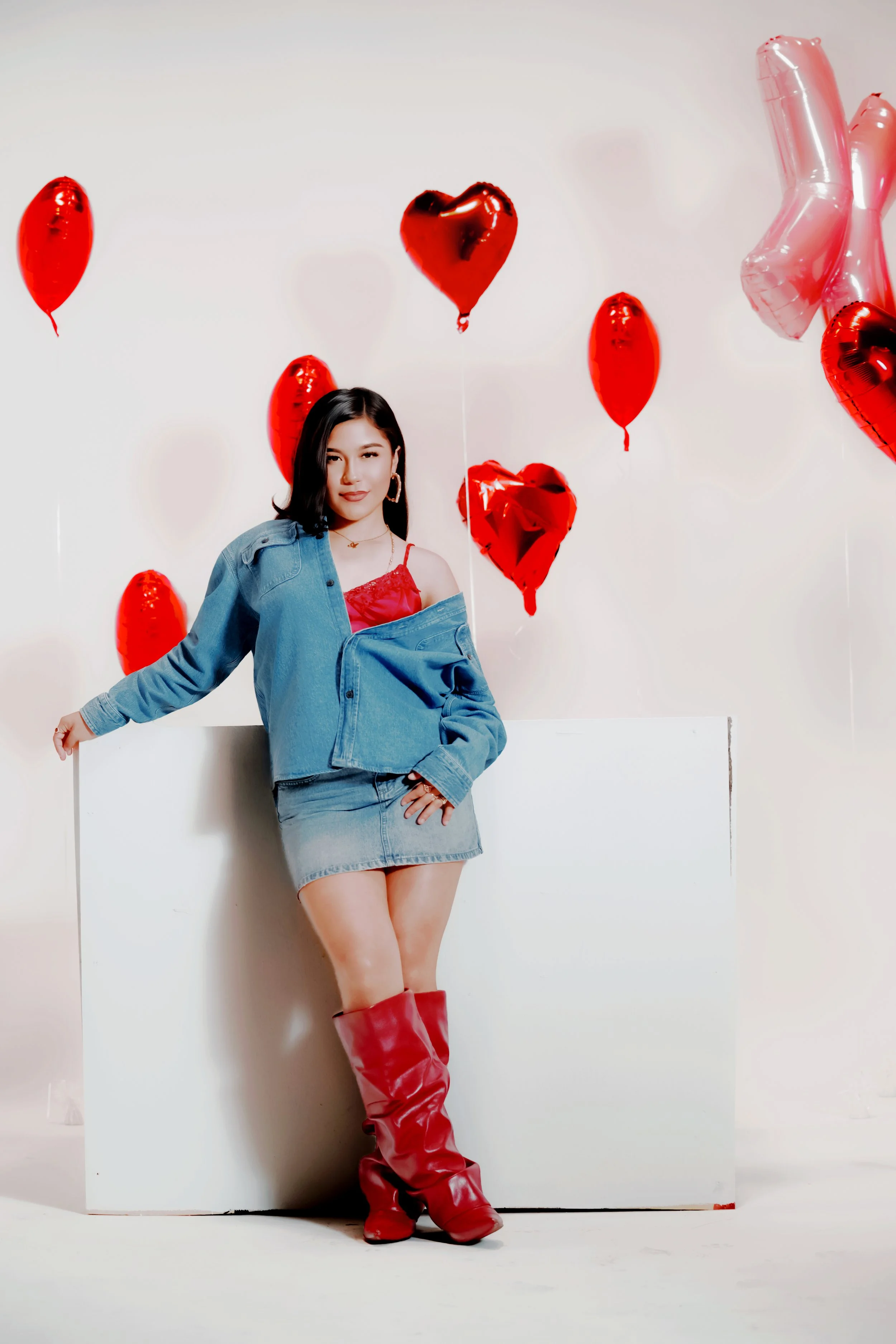 A woman dressed in a red top, denim jacket, and denim skirt, standing in front of a white wall decorated with red and pink heart-shaped balloons. She is wearing red knee-high boots and has dark hair..Hire, Book, Photographer, San Diego, Los Angeles,