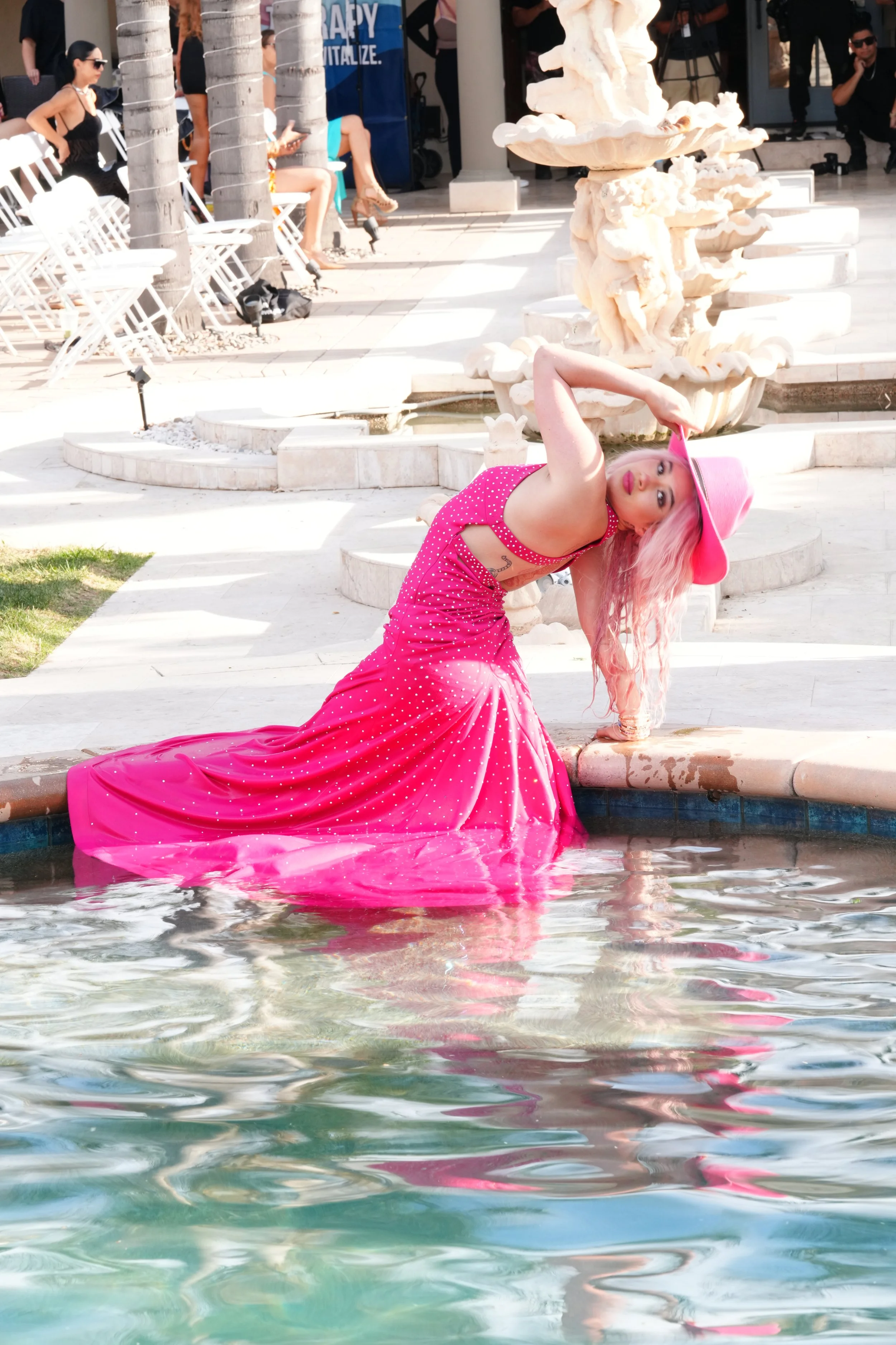 A woman with pink hair and a pink hat wearing a pink polka dot dress is posing by a fountain with water in an outdoor setting. There are people sitting on chairs and walking in the background.