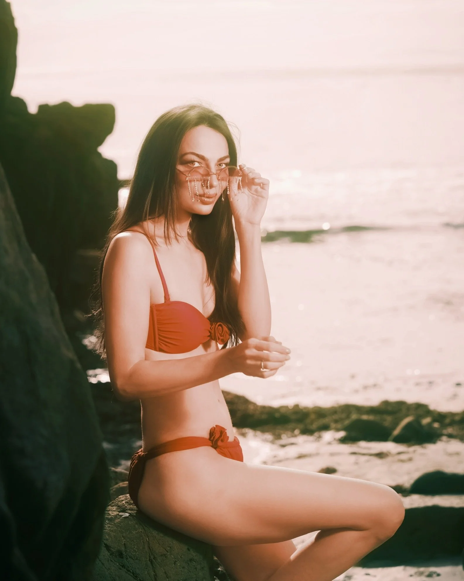 Young woman with long dark hair wearing sunglasses and a matching orange swimsuit, sitting on a rock at the beach with the ocean and sunset in the background.