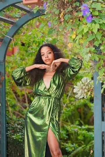 Woman in a shiny green dress standing under a garden archway with purple and white flowers and green foliage.Photographer, for hire, Oceanside, Carlsbad, San Diego, Orange County, Fashion, Portraits, Moments, Lifestyle, Events, Branding.