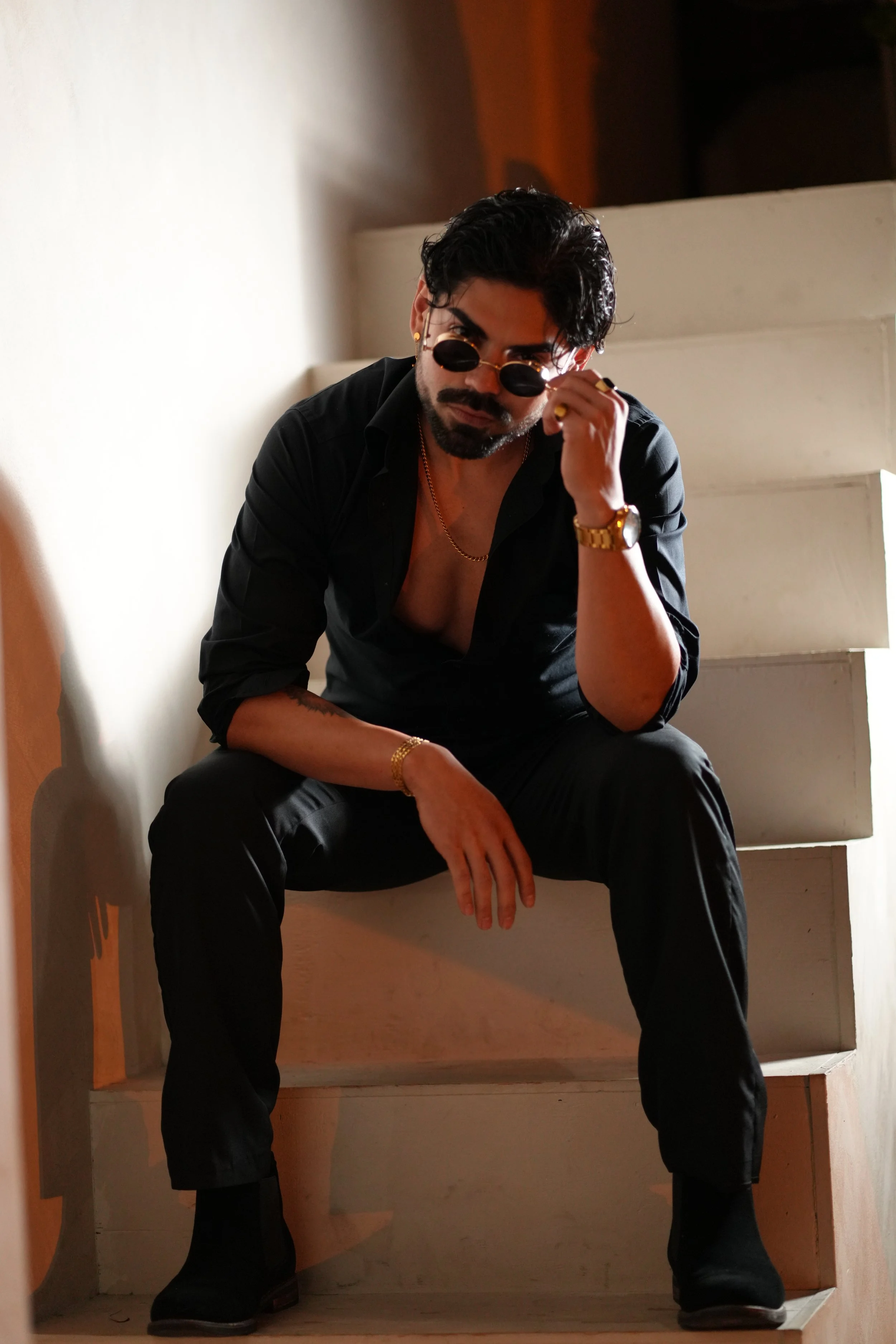A man with dark hair, sunglasses, a gold watch, and a gold chain, sitting on a staircase with a black shirt partially unbuttoned, looking at the camera with a serious expression.