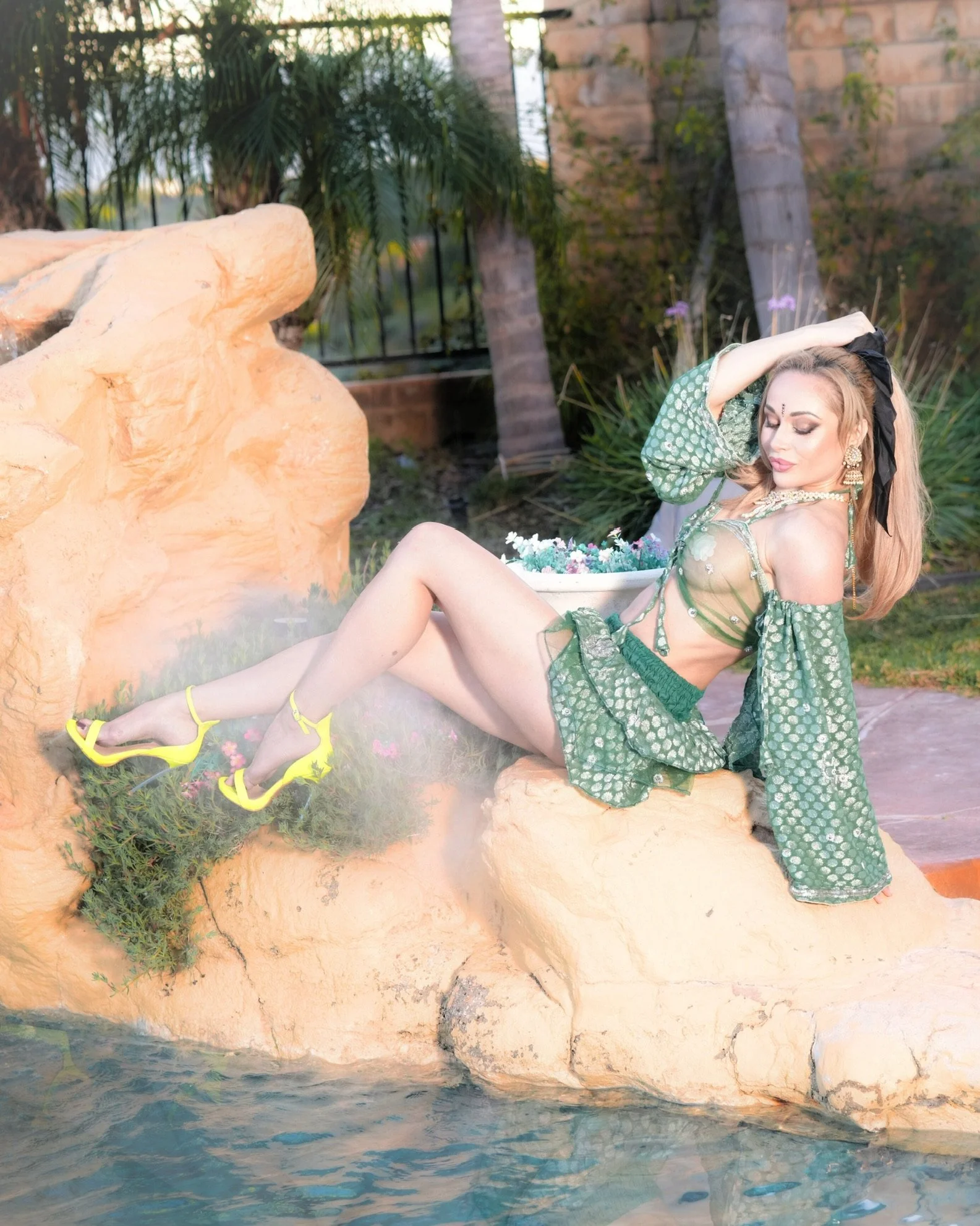A woman with long blonde hair, dressed in a green patterned outfit with yellow heels, relaxing by a pond with rocks, plants, trees, and a brick wall in the background.