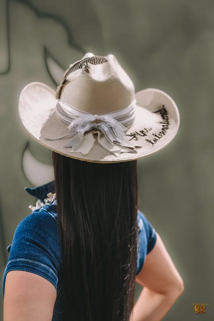 A person with long dark hair wearing a blue shirt and a decorative cowboy hat with writing on it.