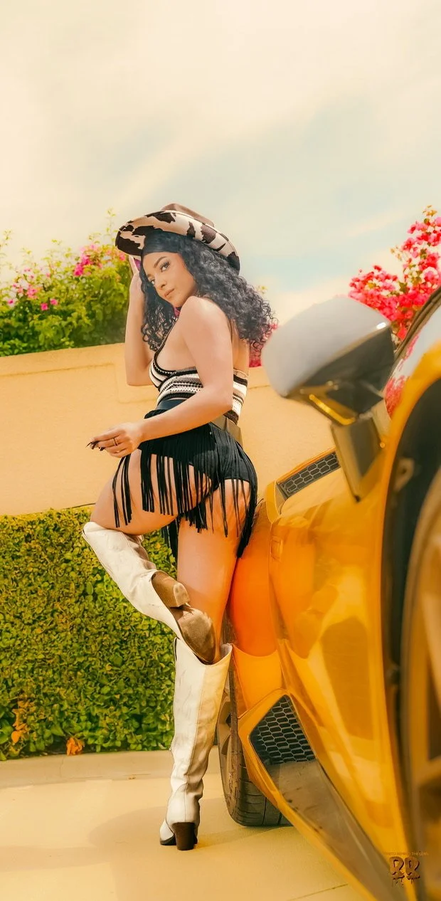 A woman with curly hair wearing a cow-print hat, a striped top, black fringed shorts, and white thigh-high boots, standing next to a yellow sports car with a background of pink flowers and bushes.