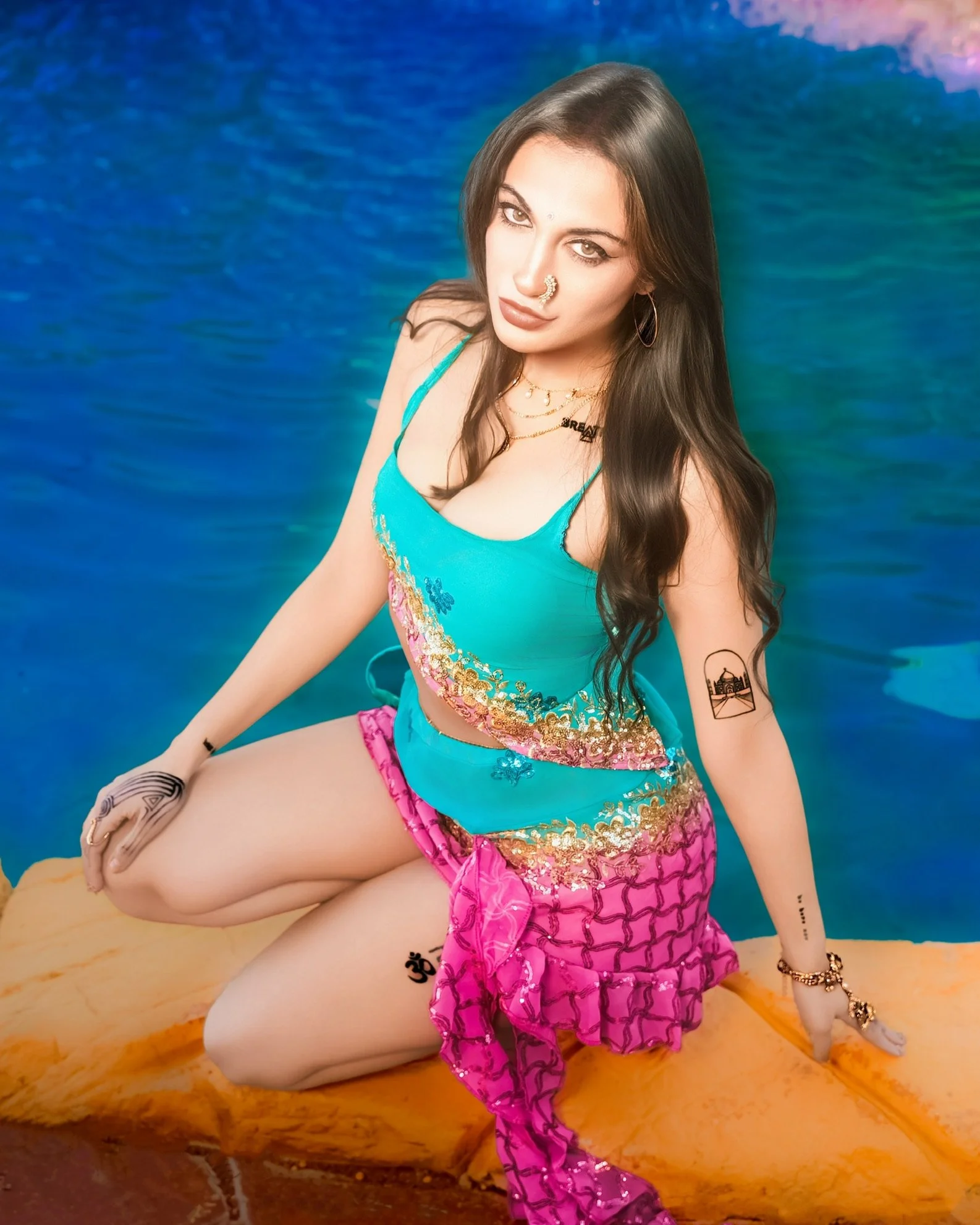 Woman with long dark hair and tattoos on her arm, wearing a colorful dress, sitting on a sandy surface with a backdrop resembling a body of water and vibrant sky colors.