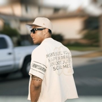 Young man wearing a white t-shirt with text on the back, a white cap, and sunglasses, walking outdoors on a street with blurred vehicles and houses in the background.Photographer, for hire, Oceanside, Carlsbad, San Diego, Orange County, Fashion, Port