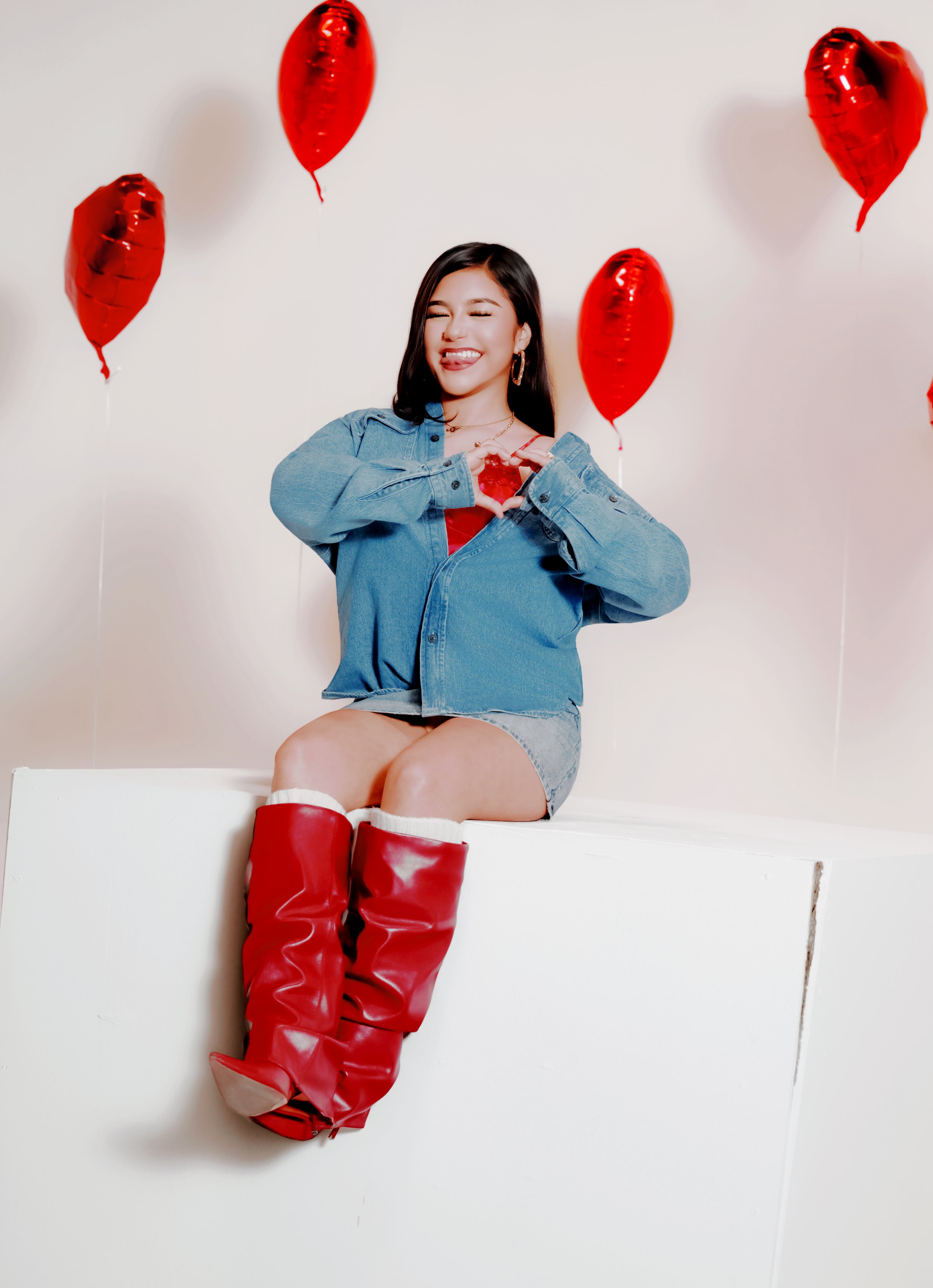A young woman with long dark hair making a face, sticking her tongue out and squinting, while forming a heart shape with her hands. She is wearing a denim jacket, gray shorts, large hoop earrings, and red knee-high boots. Red heart-shaped balloons decorate the plain white background.