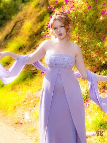 A young woman in a lavender off-the-shoulder dress standing in a garden with pink flowers and greenery, holding fabric strips in her hands.Photographer, for hire, Oceanside, Carlsbad, San Diego, Orange County, Fashion, Portraits, Moments, Lifestyle, 