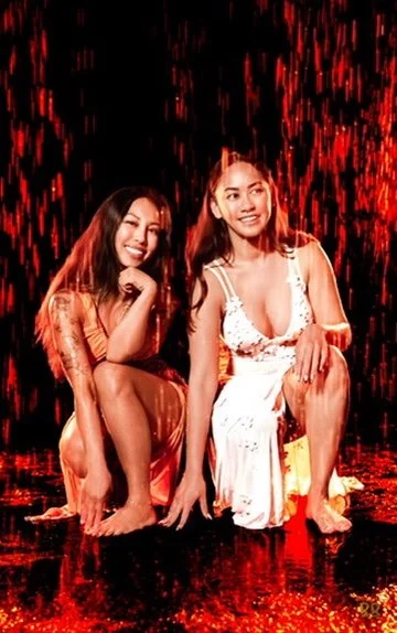 Two women squatting and smiling in a dark setting with red water or liquid falling behind them, wearing light-colored dresses.