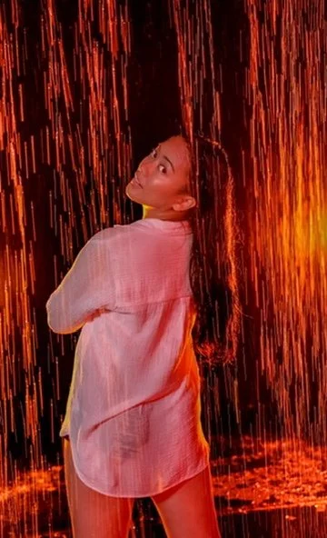 Photographer, for hire, Oceanside, Carlsbad, San Diego, Orange County, Fashion, Portraits, Moments, Lifestyle, Events, Branding.A woman stands with her back turned, looking over her shoulder, in a rain-like setting with orange and red lighting.