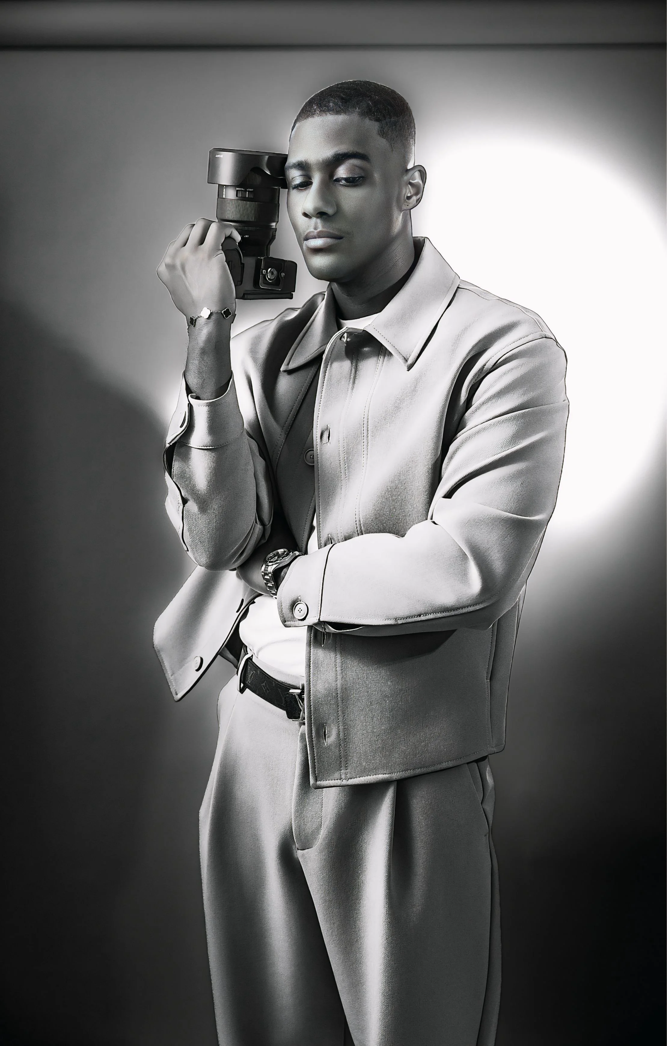 A young man with dark skin and a short haircut, wearing a light-colored suit, is holding a professional camera to his face in a studio setting. The background is plain and the lighting creates a dramatic effect.Photography Services, Modeling, Brand