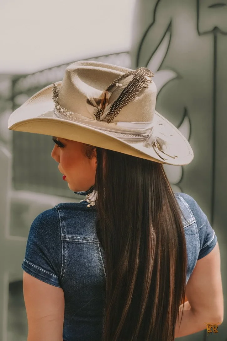 A woman wearing a large beige hat with decorative feathers and pearls, and a dark denim shirt, facing sideways with long straight hair.