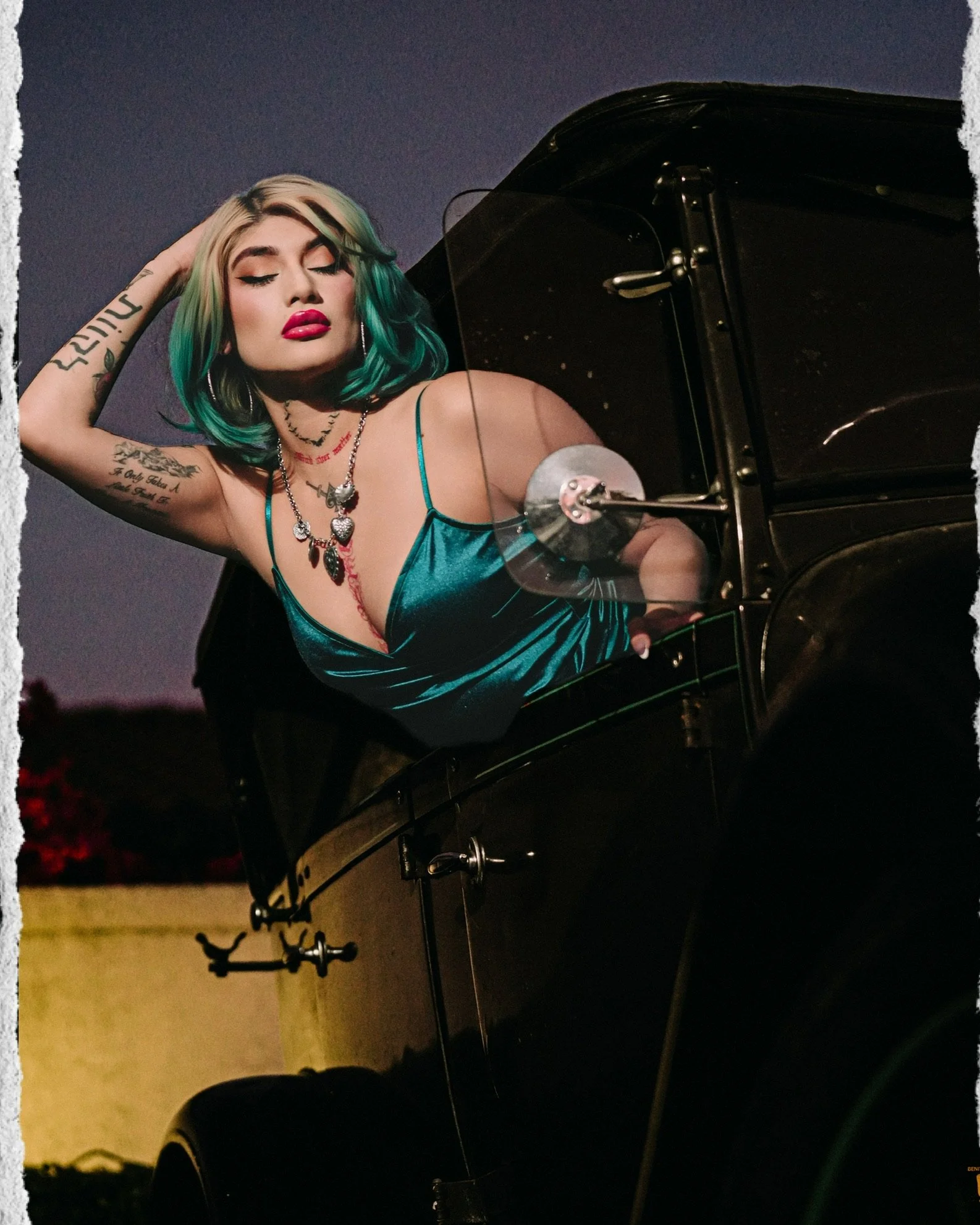 A woman with green and black hair playing a piano outdoors at dusk, wearing a teal dress, makeup with bold lips, and tattoos on her arms.