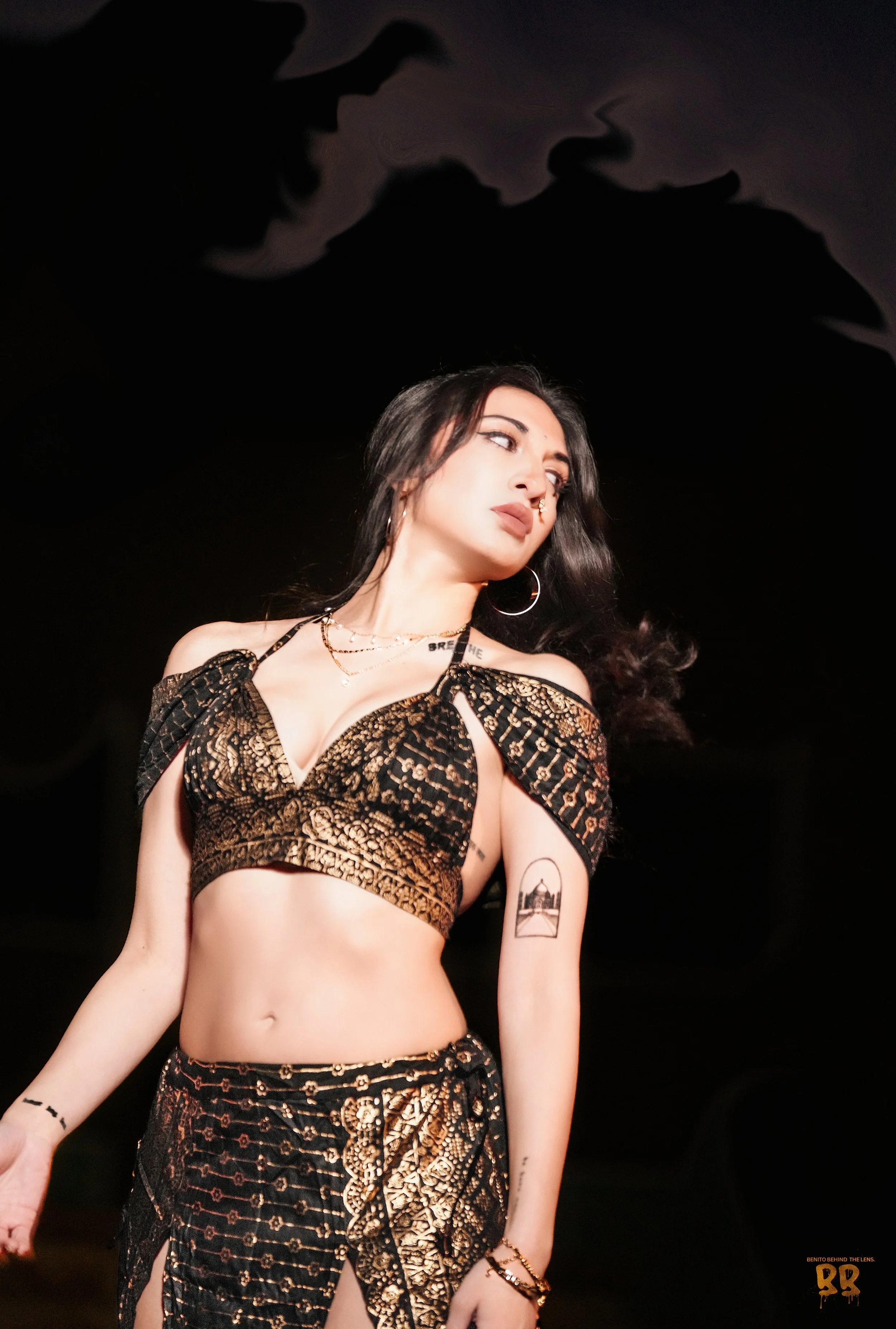 A woman with dark hair and light skin posing against a dark background. She is dressed in a black and gold patterned outfit, with visible jewelry including earrings, necklaces, and bracelets. She has tattoos on her arms and a nose ring, and appears to be looking to her right.