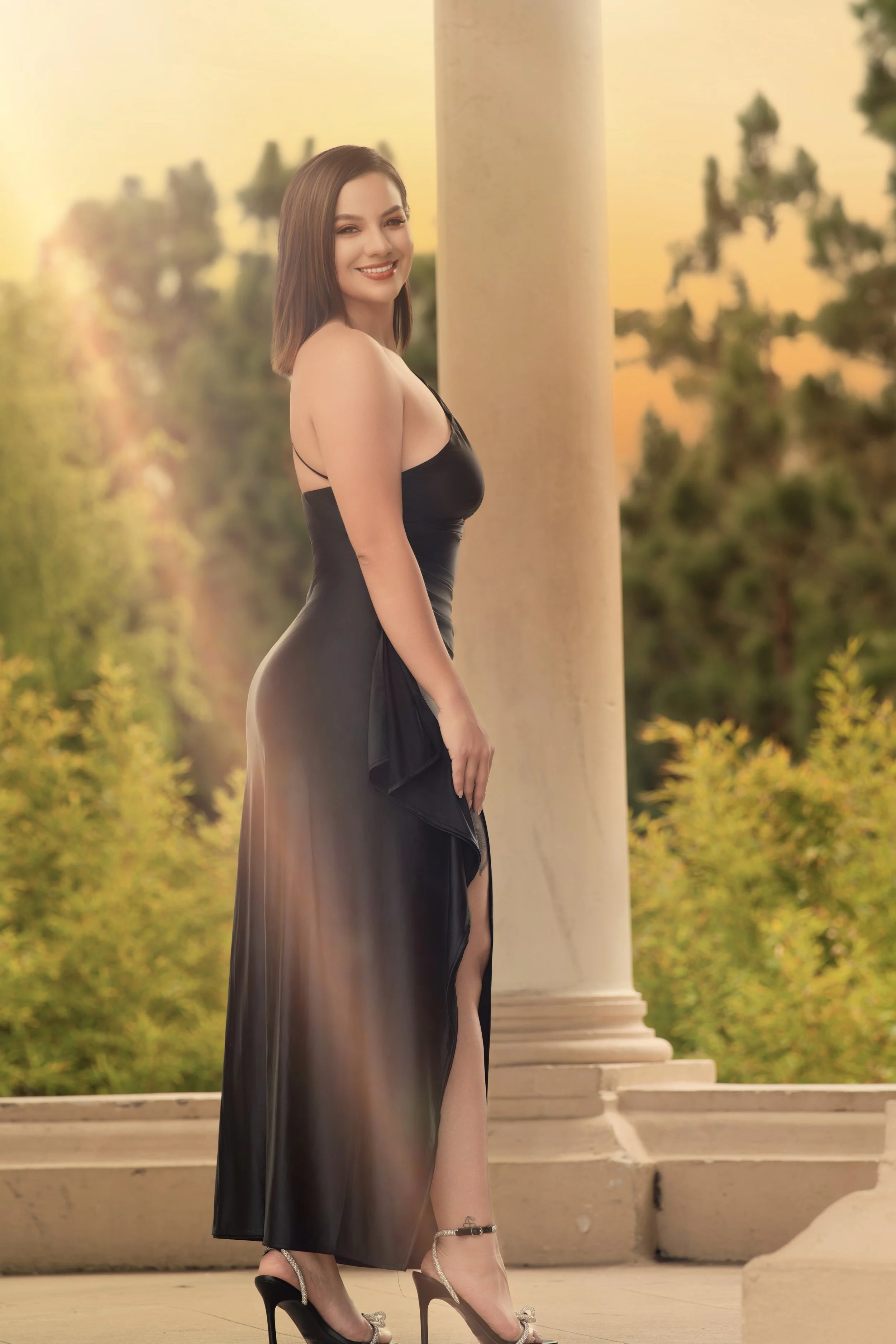 A woman in a black satin dress with a high slit, wearing high heels, stands outdoors near a cream-colored column with a sunset background.