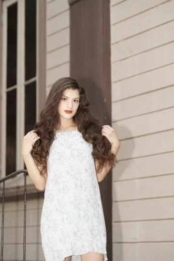 A young woman with long, wavy hair wearing a light-colored, floral dress standing outdoors near a building with wooden siding and a window.Photographer, for hire, Oceanside, Carlsbad, San Diego, Orange County, Fashion, Portraits, Moments, Lifestyle, 
