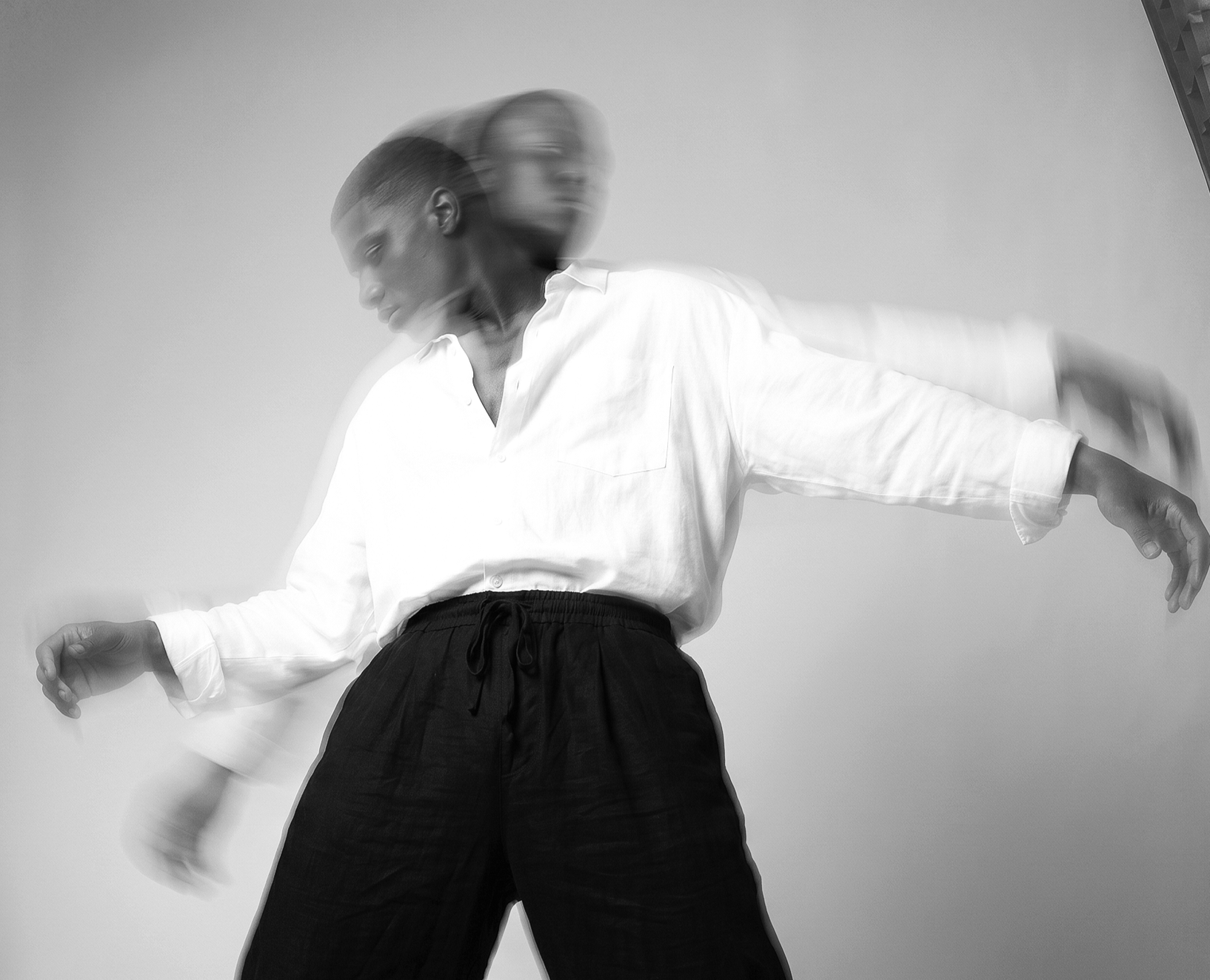 Black and white photograph of a person in motion with two overlapping faces, wearing a white shirt and dark pants, against a plain background. Photography Services, Modeling, Branding, Marketing, Fashion.
