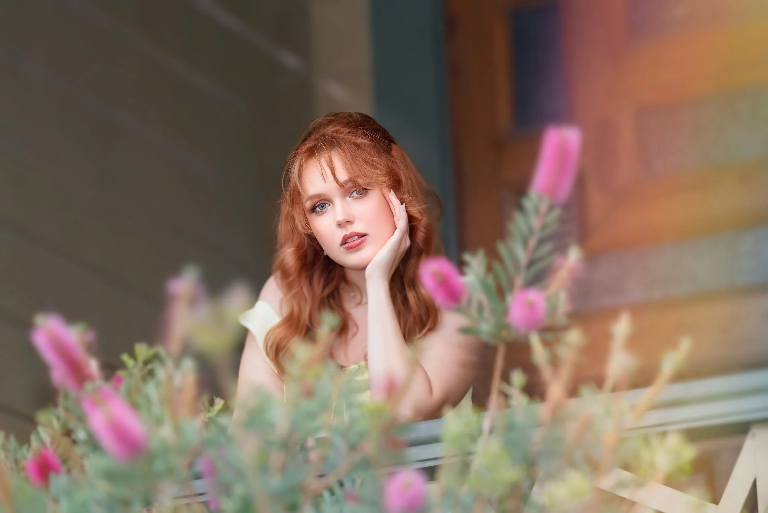 A woman with red hair and blue eyes resting her face on her hand, surrounded by pink flowers, sitting inside near a window.
