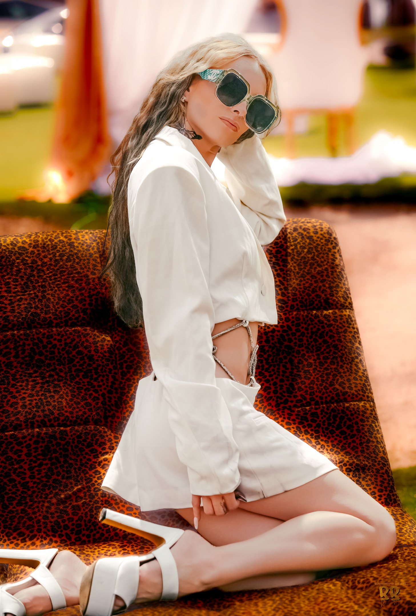 A woman with blonde and brown hair wearing oversized sunglasses, white blazer, white thigh-high stockings, and white high heels, sitting on a leopard-patterned chair in a bright room with pink walls.