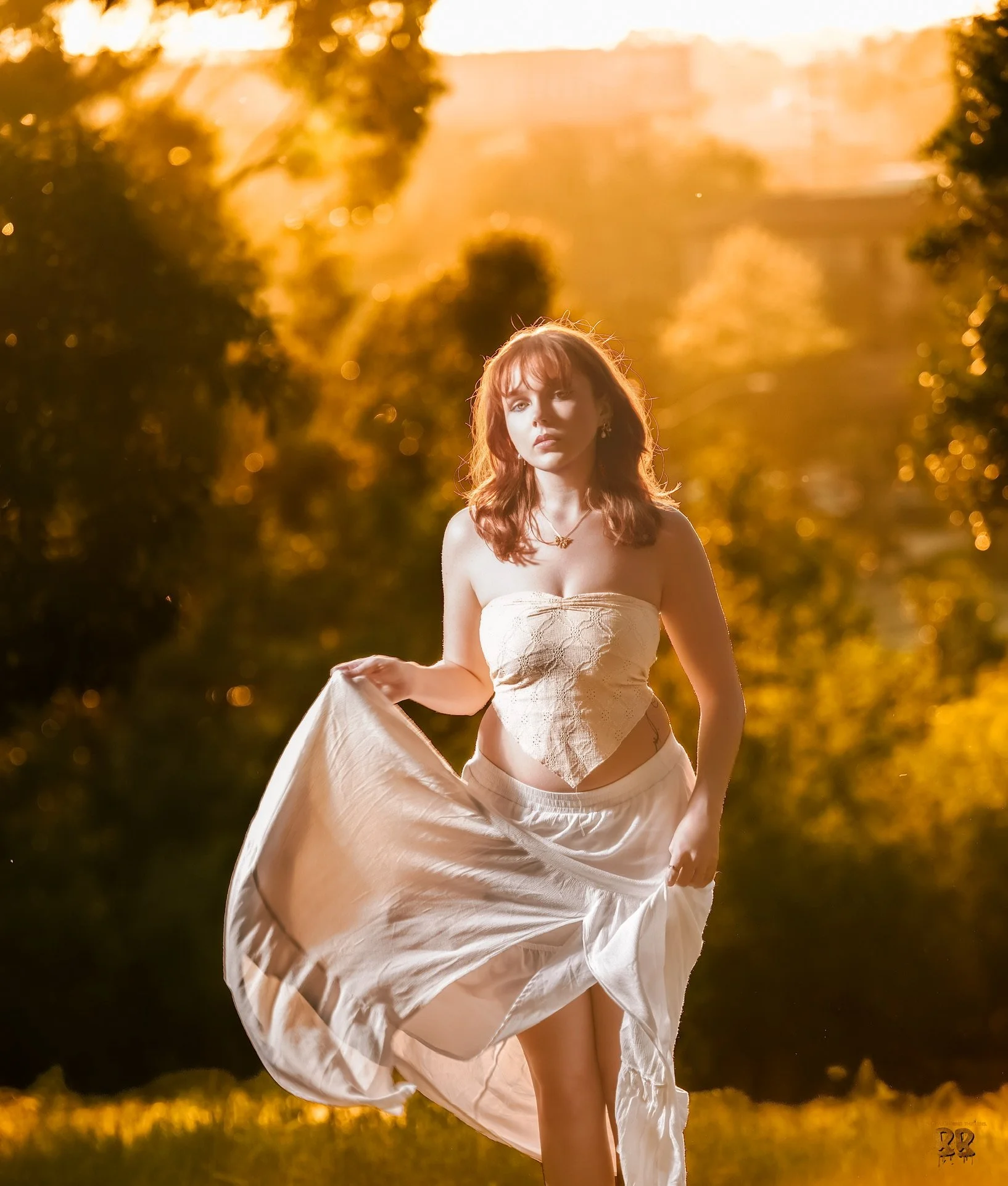 A woman with red hair stands outdoors during golden hour, holding a flowing white dress, in a setting with trees and blurred background, backlit by warm sunlight.
