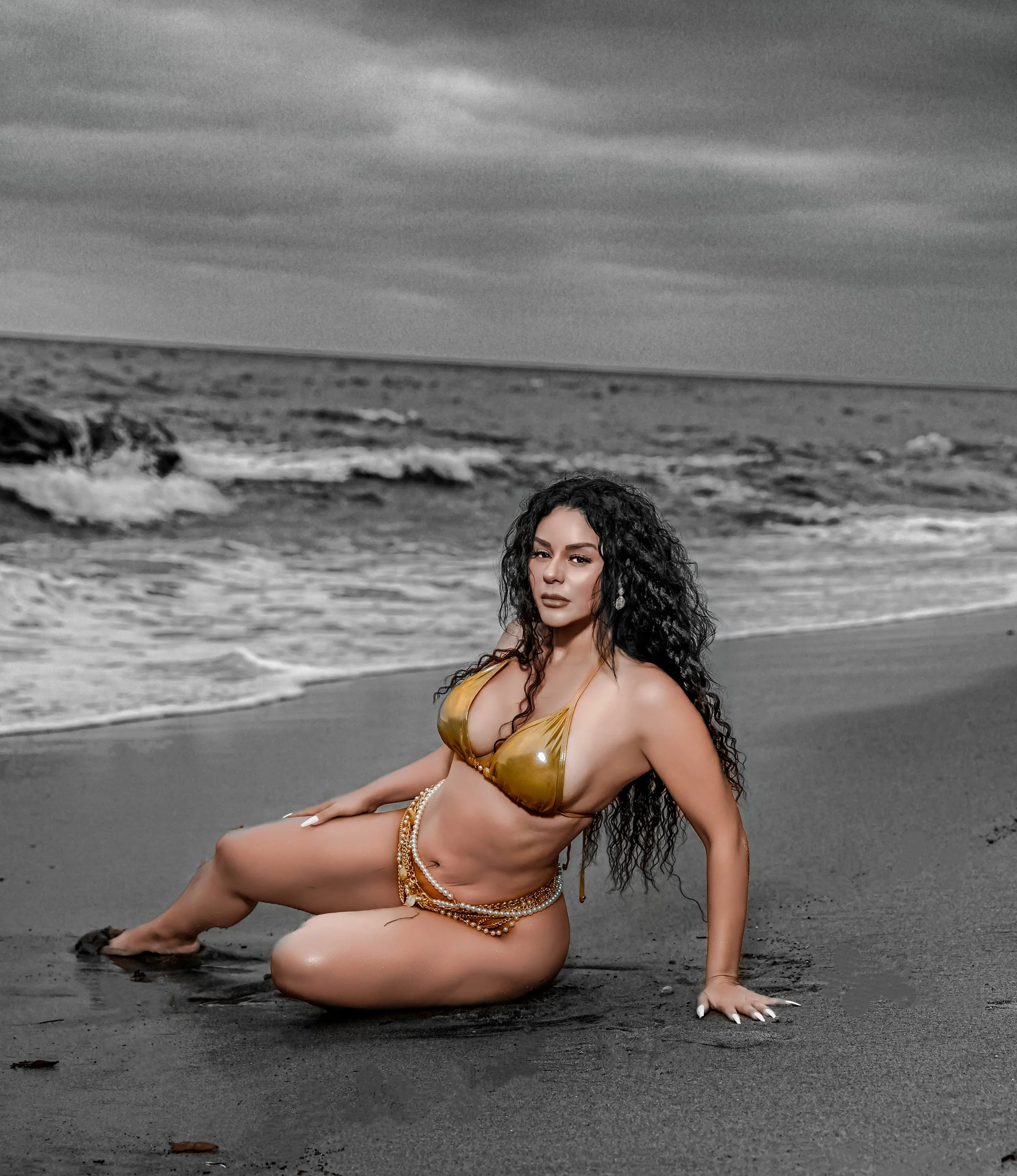 A woman with long curly dark hair and tan skin posing on a beach with the ocean in the background. She is kneeling on the wet sand, wearing a gold bikini and jewelry, looking directly at the camera with a serious expression.