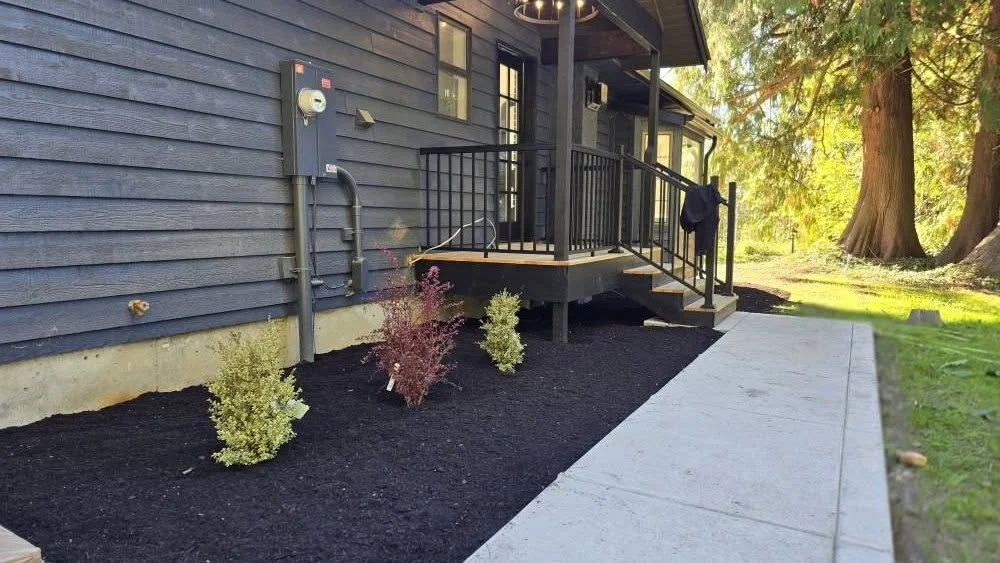 Backyard with freshly mulched flower bed, small bushes, and a concrete walkway leading to a deck with a black railing, attached to a house with dark blue exterior siding and a small window.