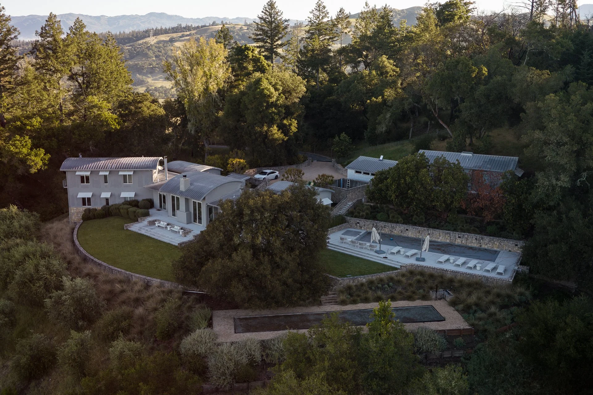 Aerial view of a modern house with a curved metal roof, large windows, and a backyard with a patio, surrounded by trees and rolling hills in the background.