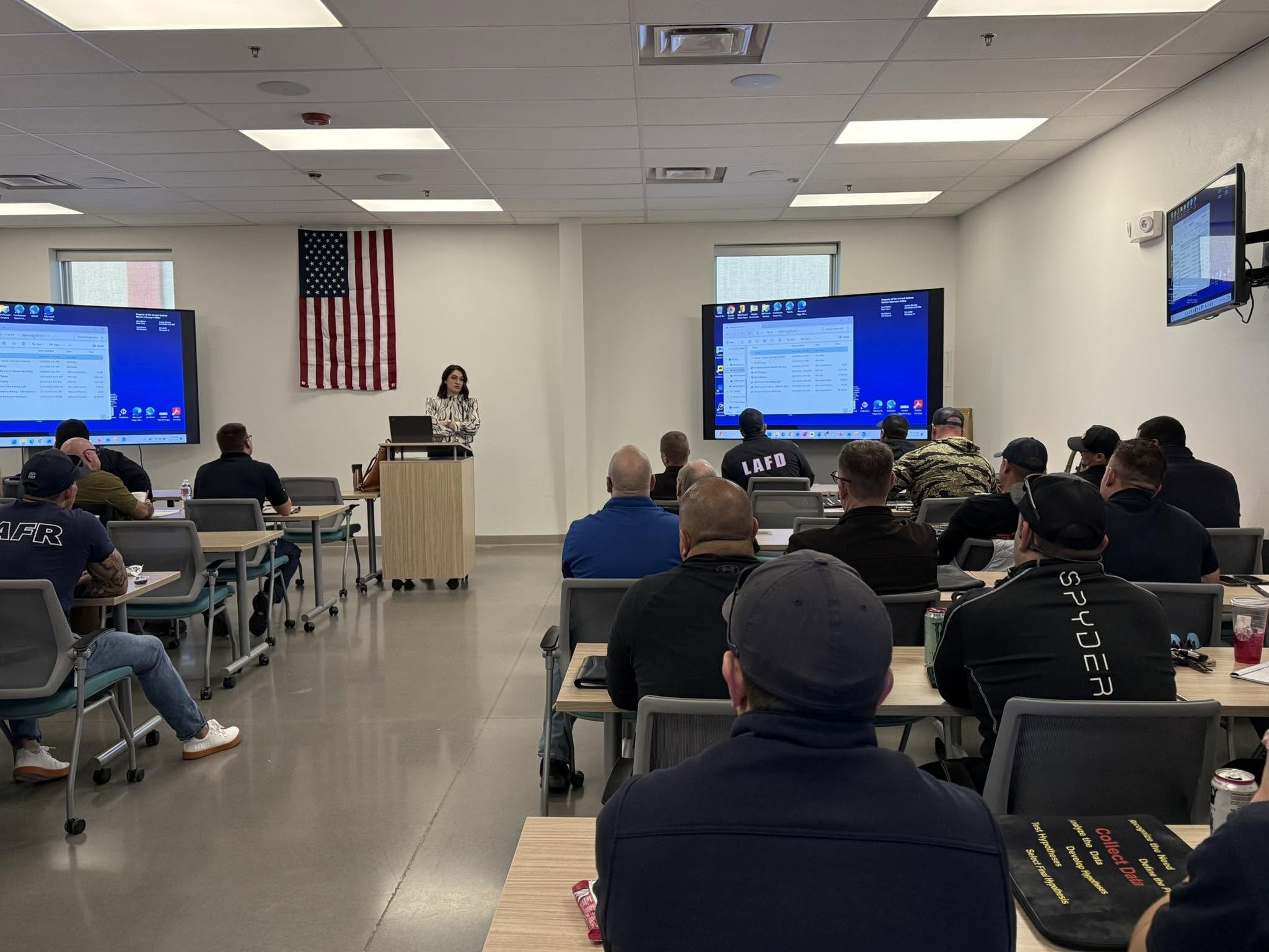 A classroom with adults attending a presentation. There is a woman standing at the front, and a large American flag mounted on the wall behind her. Students are sitting at tables, some taking notes, and there are two large screen monitors displaying 