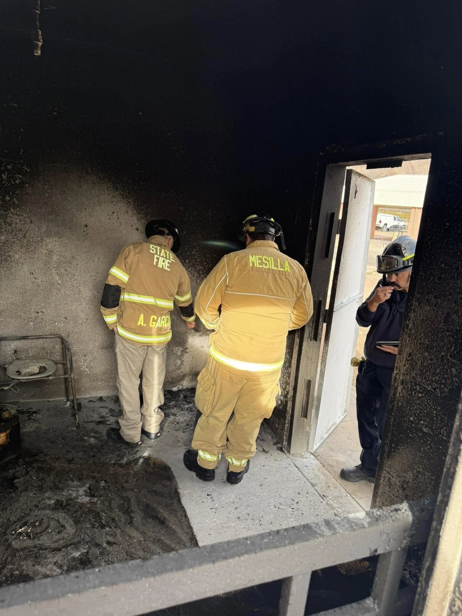 Three firefighters, two wearing yellow turnout gear with 'STATE FIRE' and 'MESILLA' written on the back, and one wearing black gear with a helmet and respirator, are inspecting the interior of a burnt building through a doorway. The interior walls ar