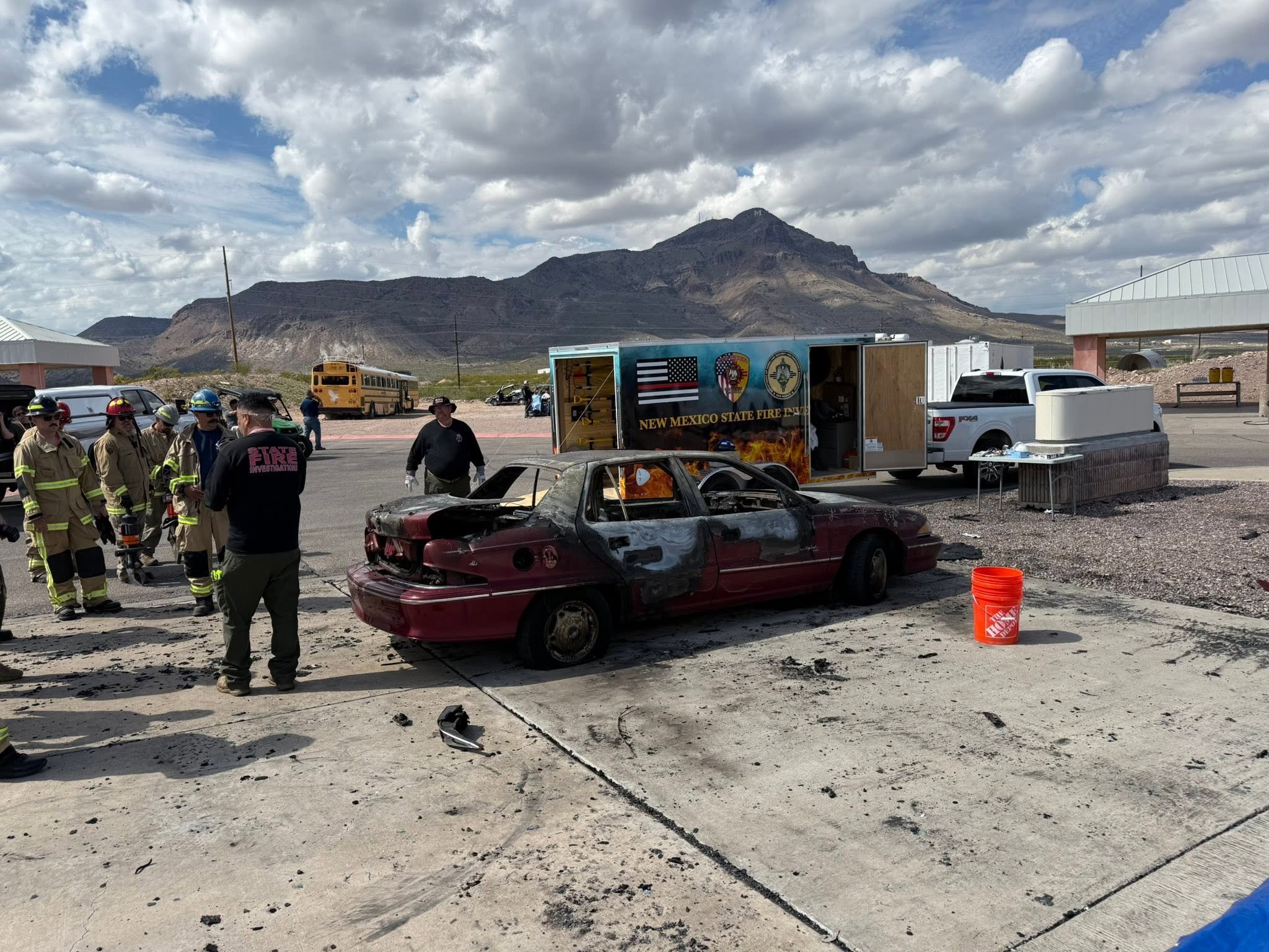 Firefighters and emergency personnel at the scene of a car fire in a parking lot with burnt vehicle and debris, with mountains and cloudy sky in the background.