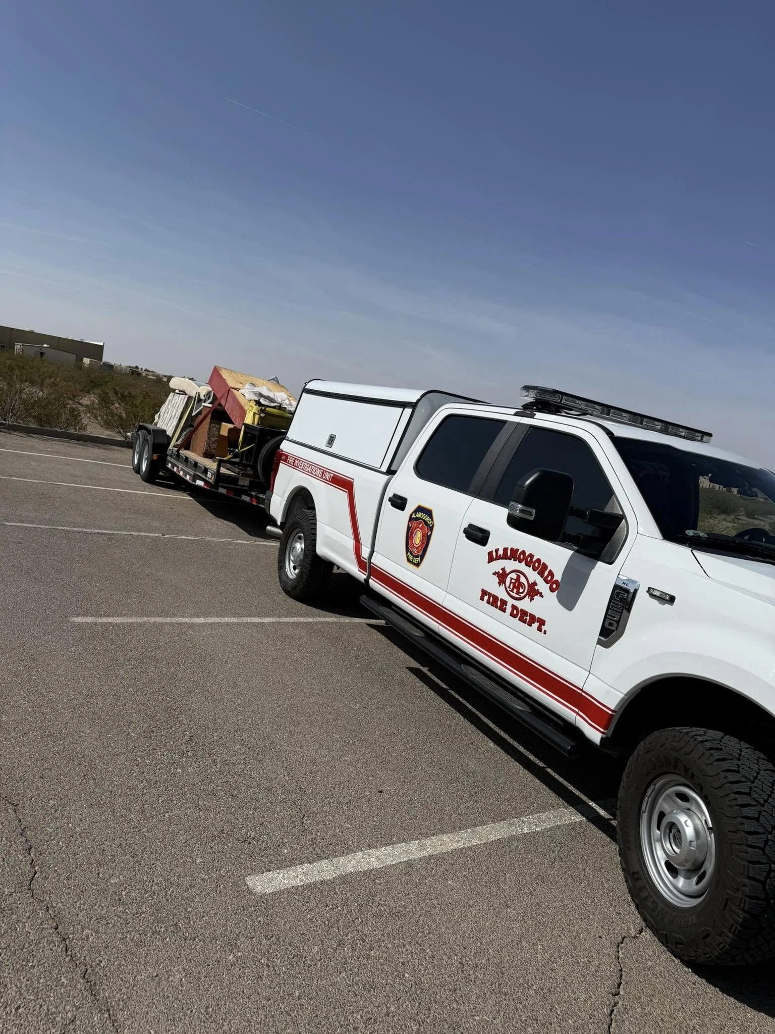 Fire department vehicle with a trailer carrying construction materials parked in an outdoor parking lot under a blue sky.
