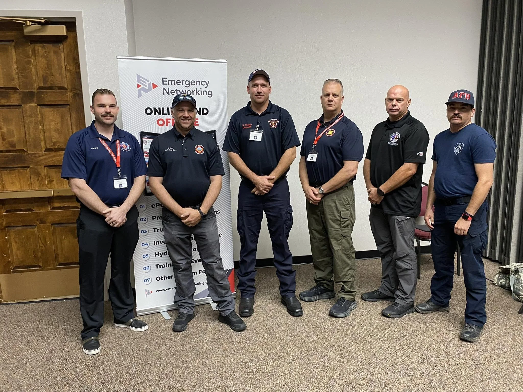 A group of six men standing indoors in front of a banner that reads 'Emergency Networking, Online and Office.' They are wearing uniforms and badges, indicating they are firefighters or emergency responders. The setting appears to be a conference or t