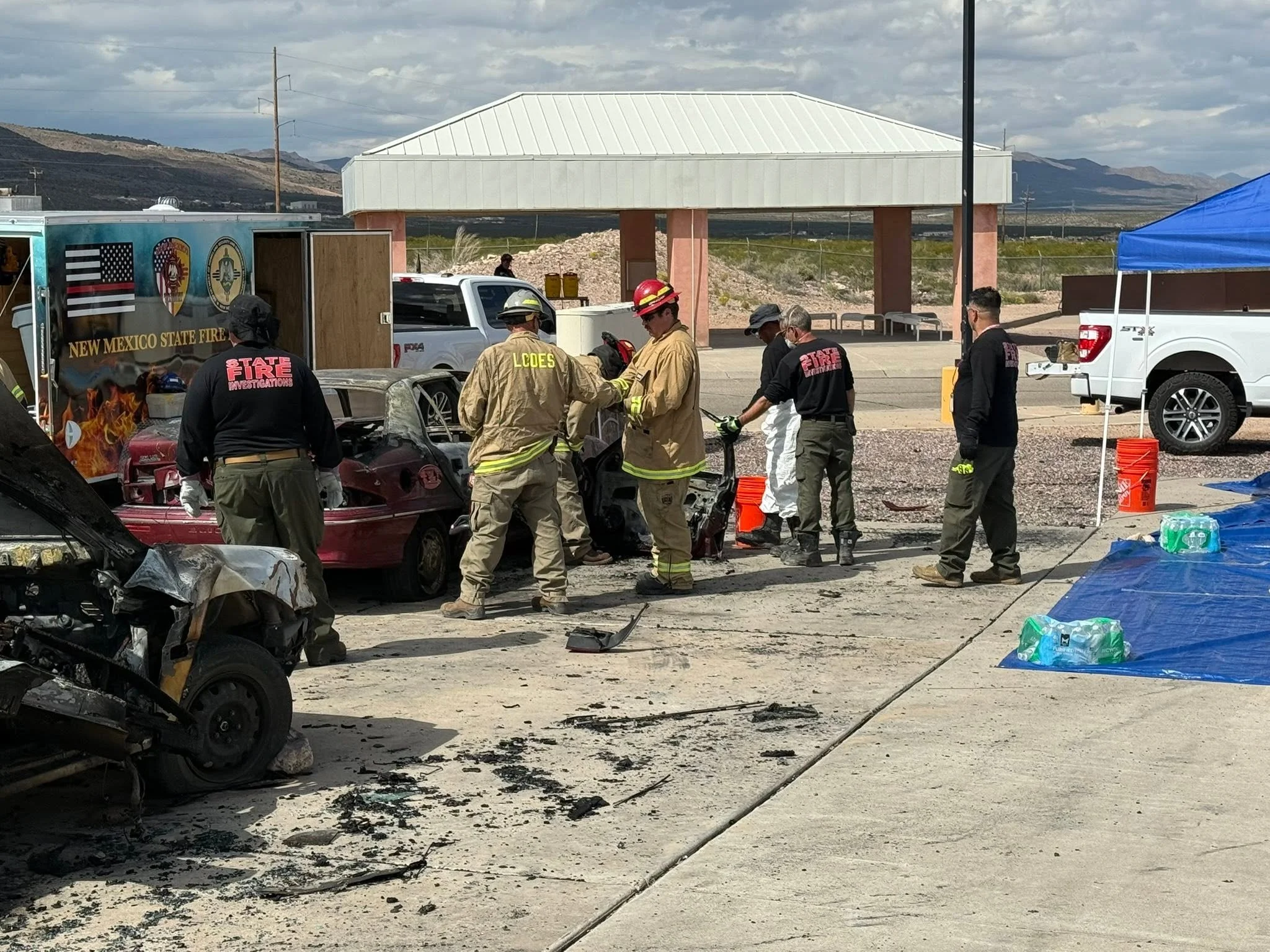 Emergency responders and firefighters at the scene of a car accident with damaged vehicles and debris, under a partly cloudy sky, in a desert-like area with mountains in the background.