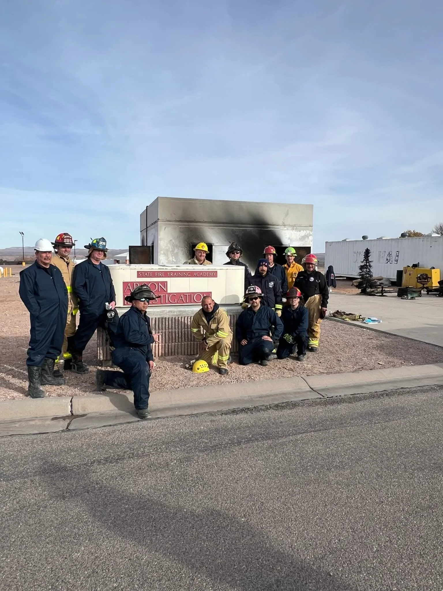Group of firefighters and emergency responders posing in front of a burnt structure with smoke damage, some in uniform with helmets, at a training academy.