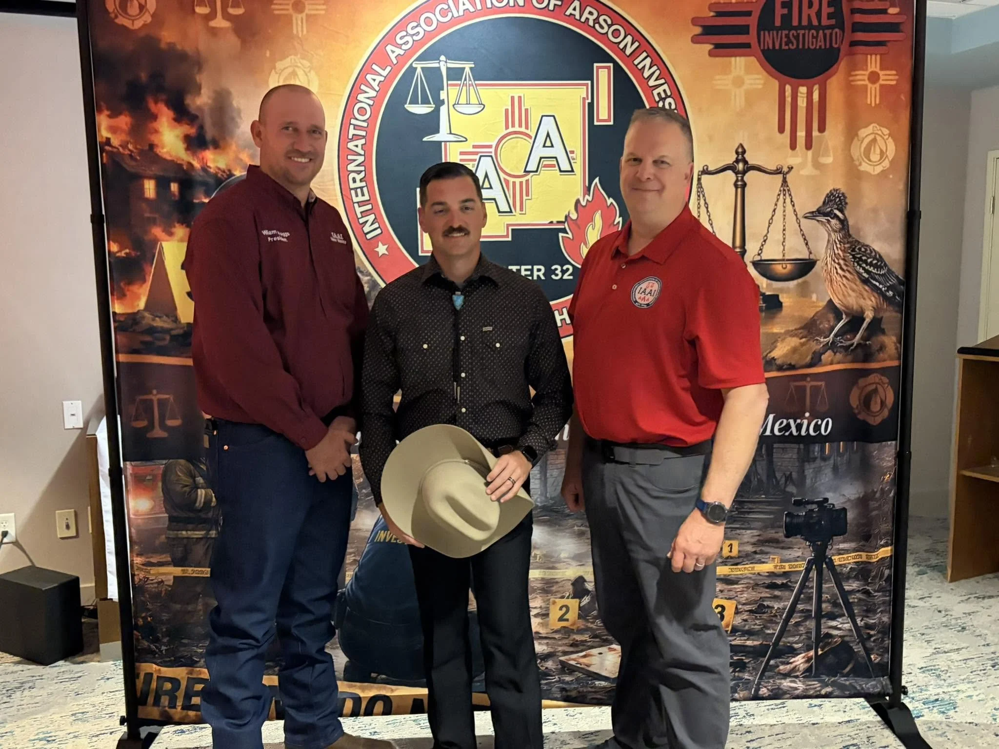 Three men standing in front of a banner with the logo of the International Association of Arson Investigators, with a background depicting a building fire, scales of justice, and a bird, with the words 'Fire Investigator' and 'Mexico'